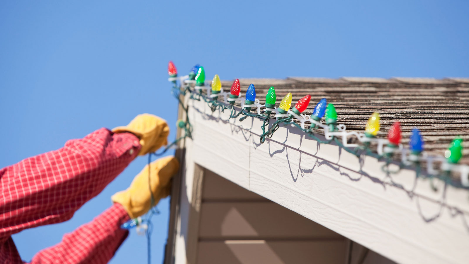 Someone wearing a red shirt and work gloves hanging holiday lights on a roof.