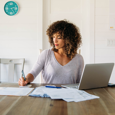 Woman at the dining room table paying papaer bills with her laptop open.
