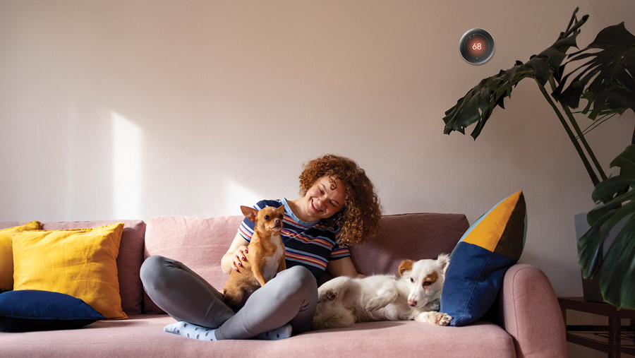 Woman sitting on her couch below a smart thermostat on the wall behind her.